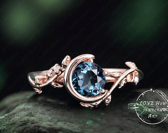 Pode incluir: Um anel de ouro rosa com uma gema de topázio azul engastada em um delicado design de folhas. O anel tem a forma de uma lua crescente e pequenos diamantes na banda.  "LOVE HOW JEWELRY ART" está impresso no canto inferior direito da imagem.