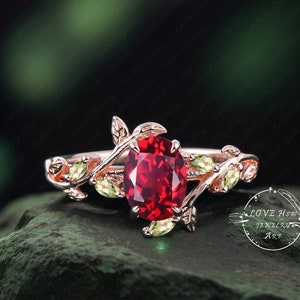 May include: A rose gold ring with a red oval gemstone surrounded by green leaves. The ring is set on a green rock.