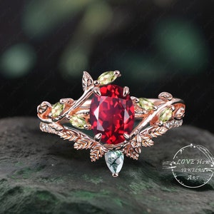 May include: Rose gold ring featuring a large oval-cut red gemstone as the centerpiece, accented with smaller green gemstones and leaf-like details. A smaller, pear-shaped green stone is set below the main stone. The ring has a nature-inspired design.