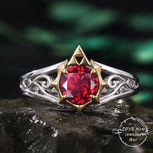 May include: A silver ring with a gold-colored setting, featuring a large, round, red gemstone. The ring has an intricate, swirling design on the band. The gemstone is set within a gold-colored frame with a triangular top.