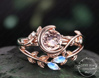 May include: Rose gold engagement ring with a delicate leaf design and a large, round, peach-colored gemstone. The ring features a crescent moon design and two smaller, pear-shaped, iridescent gemstones.