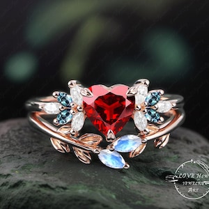 Puede incluir: Un anillo de oro rosa con una piedra roja en forma de corazón rodeada de piedras blancas y azules más pequeñas. El anillo presenta un delicado diseño de hojas con una pequeña piedra de luna en el lateral.