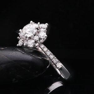 May include: A silver engagement ring with a large round center stone and smaller stones surrounding it. The band is set with smaller stones.