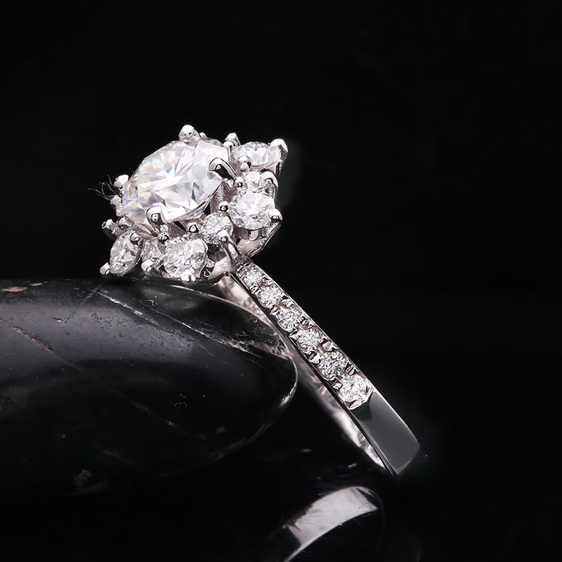 May include: A silver engagement ring with a large round center stone and smaller stones surrounding it. The band is set with smaller stones.