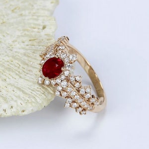 Peut inclure: Une bague en or avec une pierre précieuse en rubis rouge entourée de diamants plus petits. La bague a un design délicat en forme de feuille.