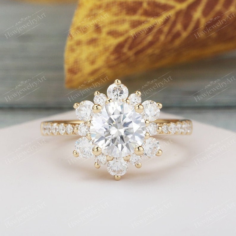 May include: A gold engagement ring with a large round center stone surrounded by a halo of smaller round stones. The ring has a delicate band with smaller stones set along the sides.