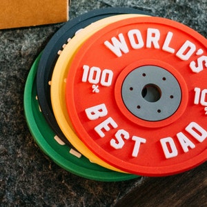 May include: Four round coasters with a weight plate design. The top coaster is red with white text that reads "WORLD'S 100% BEST DAD". The other coasters are yellow, green, and black.