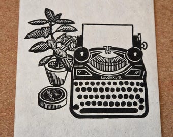 May include: Black and white linocut print featuring a vintage typewriter, a potted plant, and an ink pad. The typewriter has the text "Lousakura" on it. The print is on textured paper.
