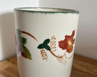 May include: A cylindrical ceramic mug with an off-white base color and a green speckled rim. The exterior features a decorative pattern of brown and green autumn leaves, including maple-like shapes and delicate brown vine tendrils.