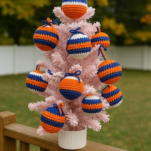 May include: A small pink Christmas tree decorated with handmade crochet ornaments. The ornaments are round, with alternating stripes of orange, blue, and white. The tree is in a white pot on a wooden deck.