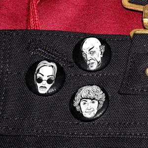 Puede incluir: Tres pines de botón en blanco y negro con diferentes caras. Los pines están unidos a una superficie de tela negra con una superficie de tela roja encima. La superficie de tela superior tiene una hebilla de metal dorado con la palabra "carhartt" en ella.