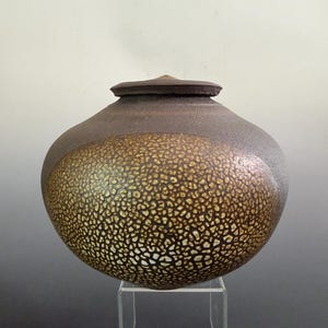 May include: A round, lidded ceramic jar with a dark, matte brown upper section and a textured, crackled golden-brown lower body. The vessel features a wide, bulbous shape that tapers toward a small opening, topped with a matching flat lid.