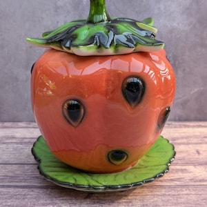 Può includere: Un contenitore in ceramica a forma di fragola con coperchio e base a forma di foglia. Il contenitore è principalmente arancione con dettagli neri e un gambo e foglie verdi. La base è verde con un bordo smerlato.