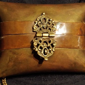May include: A small, pillow-shaped brass purse with a decorative clasp and a gold chain. The purse has two bands of copper across the front.