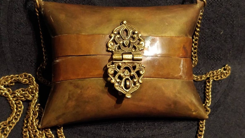 Op de afbeelding: Een klein, kussenvormig messing portemonnee met een decoratieve sluiting en een gouden ketting. De portemonnee heeft twee banden van koper aan de voorkant.