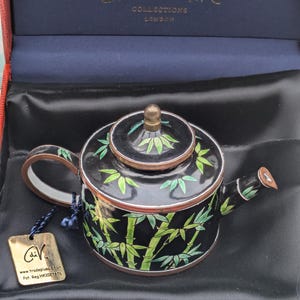 Könnte beinhalten: Eine kleine schwarze und grüne Teekanne mit Bambus-Design. Die Teekanne befindet sich in einer roten Schachtel mit blauer Auskleidung. Die Schachtel trägt den Text "Charlotte di Vita" und "Collections London".