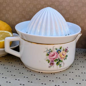 May include: White ceramic citrus juicer with a floral design and gold trim. The juicer has a reamer and a bowl with a handle. A lemon is visible in the background. Kitchenware.
