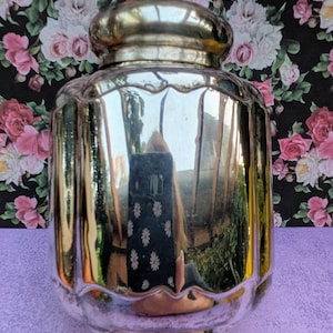 Könnte beinhalten: Ein Vintage-Glasbehälter mit Spiegelglas und einem goldfarbenen Deckel. Der Behälter hat einen abgerundeten Boden und ein gewelltes Design in der Mitte. Die reflektierende Oberfläche spiegelt die Umgebung wider. Der Hintergrund zeigt ein Blumenmuster.