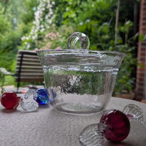 Puede incluir: Un cuenco de cristal transparente con superficie texturizada y una pequeña asa en la tapa. Tres pequeños caramelos de cristal con alas transparentes están colocados alrededor del cuenco, con un centro de color rojo, azul y rosa oscuro.