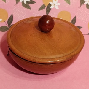 Könnte beinhalten: Eine braune Holzschale mit Deckel und einem Holzgriff oben. Die Schale steht auf einem rosa Hintergrund mit einem Blumenmuster.