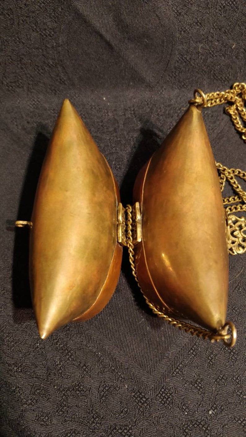 Op de afbeelding: Een messing scharnierende tas met een ketting eraan. De tas is open en de binnenkant is zichtbaar. De tas is traanvormig.