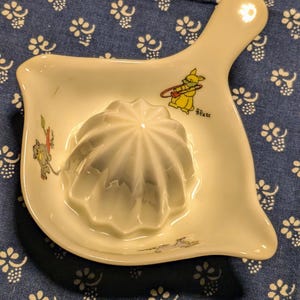 May include: White ceramic citrus juicer with a handle and a small, stylized illustration of a rabbit playing a musical instrument. The juicer has a fluted cone and a small, flat base. The illustration is in yellow, red, and blue.