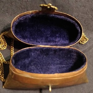 Op de afbeelding: Een klein, ovaalvormig, messing tasje met een gouden ketting. Het tasje is open, waardoor een blauwe fluwelen voering zichtbaar is.