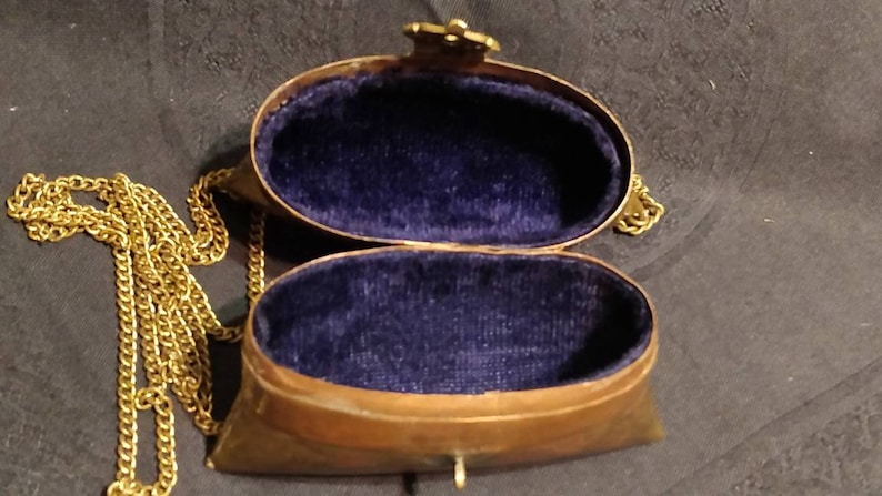 Op de afbeelding: Een klein, ovaalvormig, messing tasje met een gouden ketting. Het tasje is open, waardoor een blauwe fluwelen voering zichtbaar is.