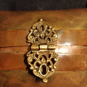 Op de afbeelding: Een klein, rechthoekig doosje met een scharnierend deksel en een decoratieve messing sluiting. Het doosje is gemaakt van koper en heeft een gelaagde, gestreepte uitstraling. Een gouden ketting is aan het doosje bevestigd.
