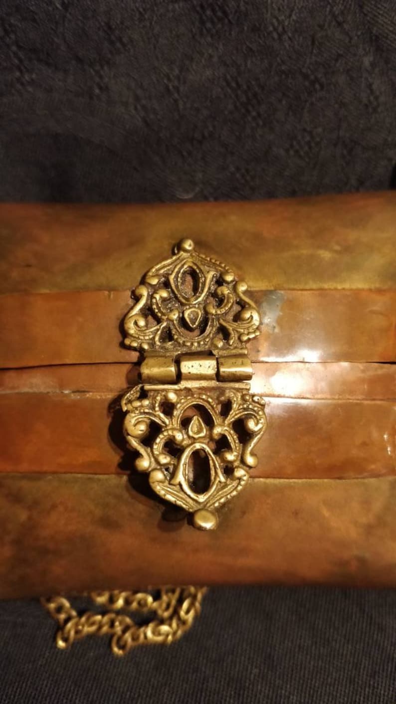 Op de afbeelding: Een klein, rechthoekig doosje met een scharnierend deksel en een decoratieve messing sluiting. Het doosje is gemaakt van koper en heeft een gelaagde, gestreepte uitstraling. Een gouden ketting is aan het doosje bevestigd.