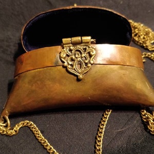 Op de afbeelding: Een klein, messingkleurig, scharnierdoosje met een ketting. Het doosje is open en de binnenkant is bekleed met blauw fluweel. Het doosje heeft een decoratieve, sierlijke messing sluiting.