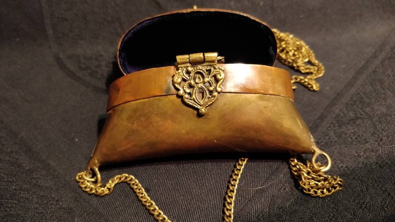 Op de afbeelding: Een klein, messingkleurig, scharnierdoosje met een ketting. Het doosje is open en de binnenkant is bekleed met blauw fluweel. Het doosje heeft een decoratieve, sierlijke messing sluiting.