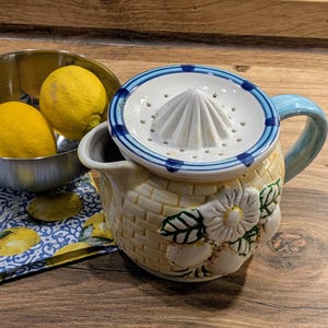 Könnte beinhalten: Ein gelber Keramik-Krug mit blau-weißem Rand und einem weißen Keramikdeckel mit einem Sieb. Der Krug hat ein Blumenmuster und ein Korbgeflechtmuster. Der Deckel hat einen weißen Keramikkegel in der Mitte.