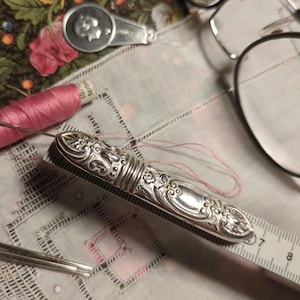 Op de afbeelding: Een antiek zilveren naaldenkoker met sierlijke bloemendetails, rustend op een liniaal. Een spoel roze garen en een bril zijn ook zichtbaar. De naaldenkoker is ongeveer 7.6 cm lang en de afbeelding suggereert een naai- of knutselthema.