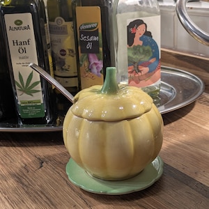 Può includere: Una zuccheriera in ceramica gialla a forma di zucca con gambo verde e piattino abbinato. Un cucchiaino è all'interno. Sullo sfondo ci sono diverse bottiglie di olio.