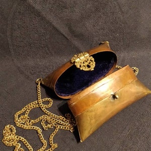 Op de afbeelding: Een klein, ovaalvormig messing portemonnee met een gouden kettingband. De portemonnee is open, waardoor een blauwe fluwelen voering zichtbaar is. De portemonnee heeft een decoratieve messing sluiting.