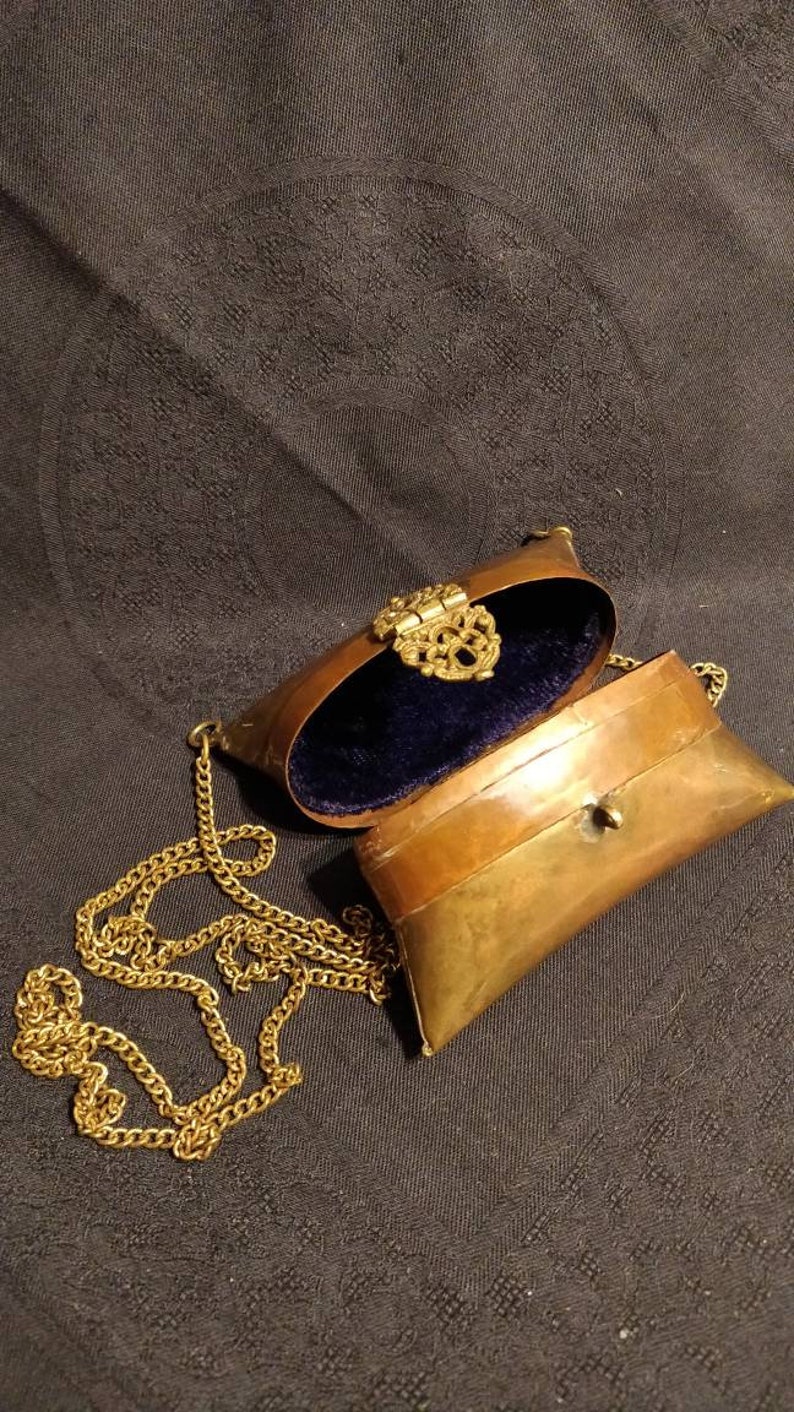 Op de afbeelding: Een klein, ovaalvormig messing portemonnee met een gouden kettingband. De portemonnee is open, waardoor een blauwe fluwelen voering zichtbaar is. De portemonnee heeft een decoratieve messing sluiting.