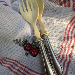 Könnte beinhalten: Ein Set aus zwei Salatbesteck mit elfenbeinfarbenen Griffen und versilberten Zinken. Das Besteck liegt auf einem weißen und rot gestreiften Küchentuch.