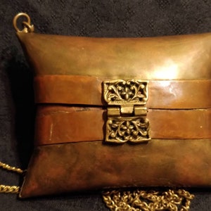 Op de afbeelding: Een kussenvormige handtas van messing en koper met een decoratieve sluiting en een gouden kettingband.