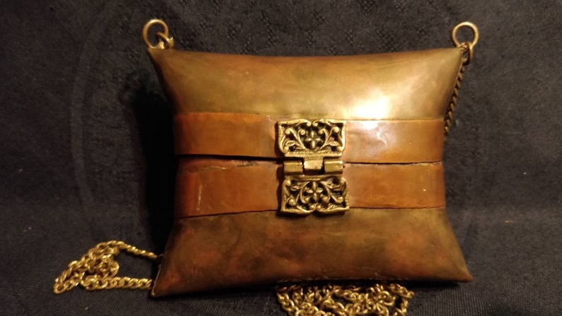 Op de afbeelding: Een kussenvormige handtas van messing en koper met een decoratieve sluiting en een gouden kettingband.