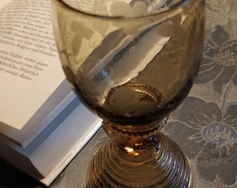 May include: A brown glass goblet with a decorative design and a wide, flared base. The goblet is sitting on a patterned surface.