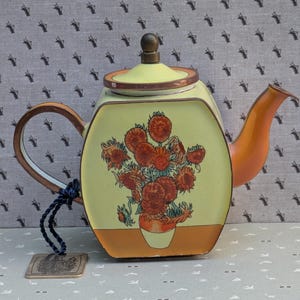 May include: A light green, hexagonal metal teapot featuring a painted illustration of orange sunflowers in a vase on the front. The teapot has a copper-colored handle, spout, and lid rim, with a dark blue cord attached to a metal tag at the base.