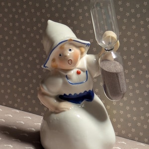 May include: A ceramic figurine of a Dutch girl holding a clear hourglass. The figurine is dressed in a white outfit with blue and red accents. The hourglass contains dark sand. The figurine has a surprised expression.