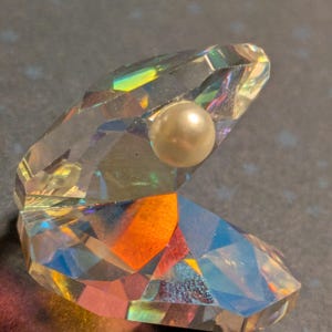 Puede incluir: Una concha de cristal transparente y facetada con una perla en su interior. El cristal refleja un arcoíris de colores, incluyendo naranja, amarillo y azul. La perla es de color blanco crema. La concha está abierta, mostrando la perla.