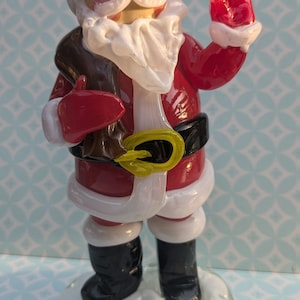 Puede incluir: Una figurita de vidrio de Papá Noel con un traje rojo con ribetes blancos, una barba blanca y botas negras. Lleva una bolsa marrón en una mano y saluda con la otra. Está de pie sobre una base blanca.