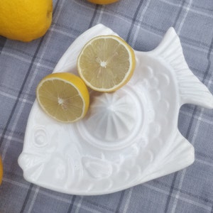 May include: White ceramic fish-shaped citrus juicer with two lemon halves on top. The juicer has a textured surface and a raised design.