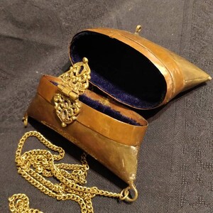 Op de afbeelding: Een klein, messingkleurig, scharnierdoosje met een ketting eraan. Het doosje is open en bekleed met blauw fluweel. Het doosje heeft een sierlijke messing sluiting.