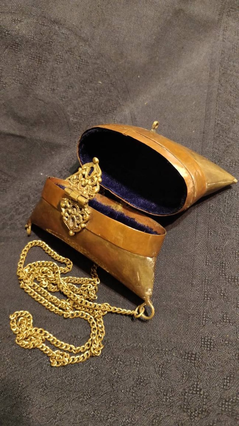 Op de afbeelding: Een klein, messingkleurig, scharnierdoosje met een ketting eraan. Het doosje is open en bekleed met blauw fluweel. Het doosje heeft een sierlijke messing sluiting.