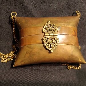 Op de afbeelding: Een kussenvormige portemonnee van messing met een scharnierende sluiting en een gouden ketting.