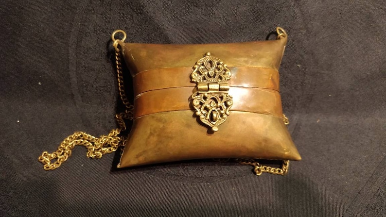 Op de afbeelding: Een kussenvormige portemonnee van messing met een scharnierende sluiting en een gouden ketting.